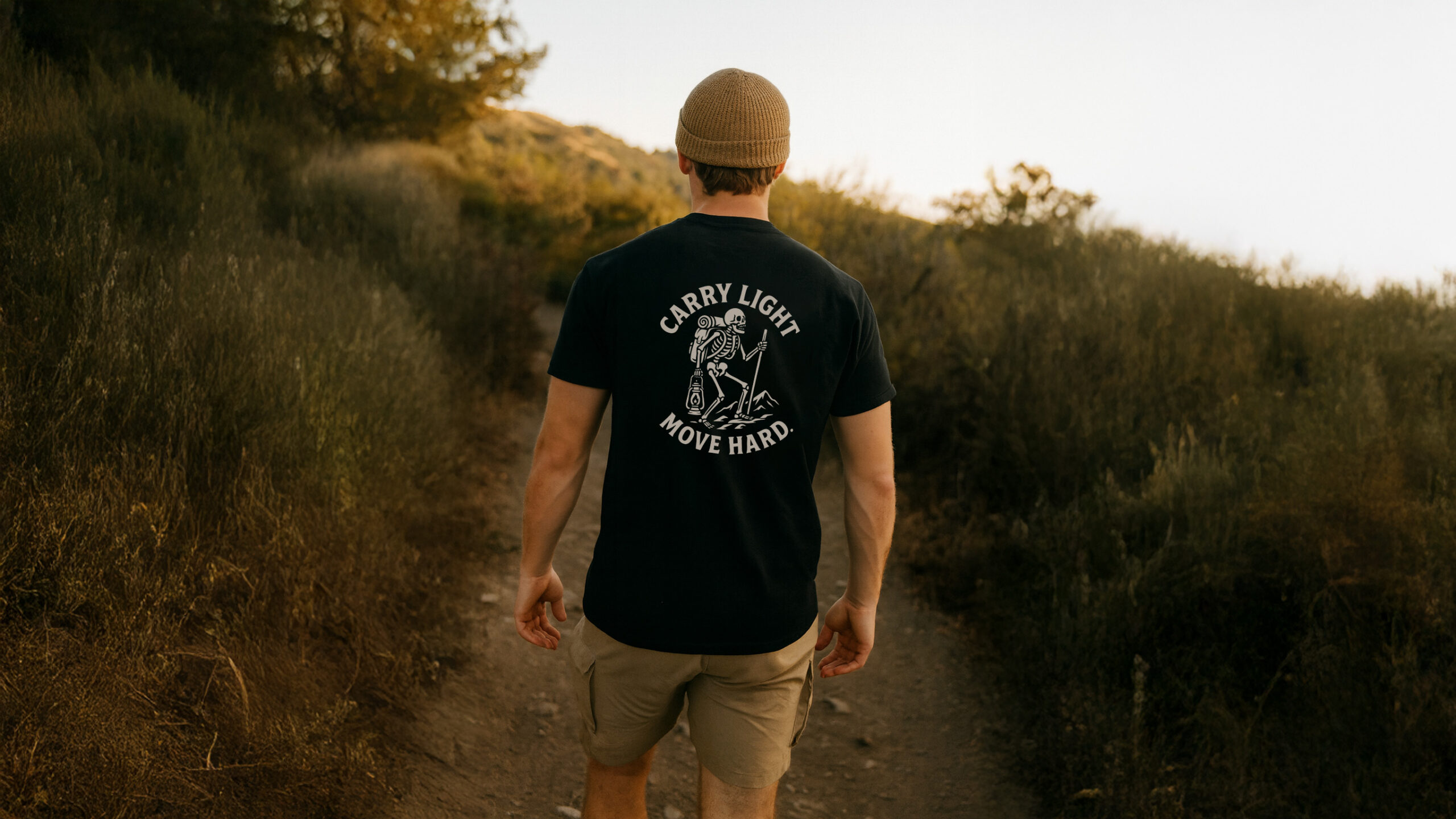 Man hiking a dirt trail at sunset wearing the Varbridge “Carry Light, Move Hard” black graphic tee with skeletal explorer design - rugged workwear-inspired t-shirt for adventure and endurance.