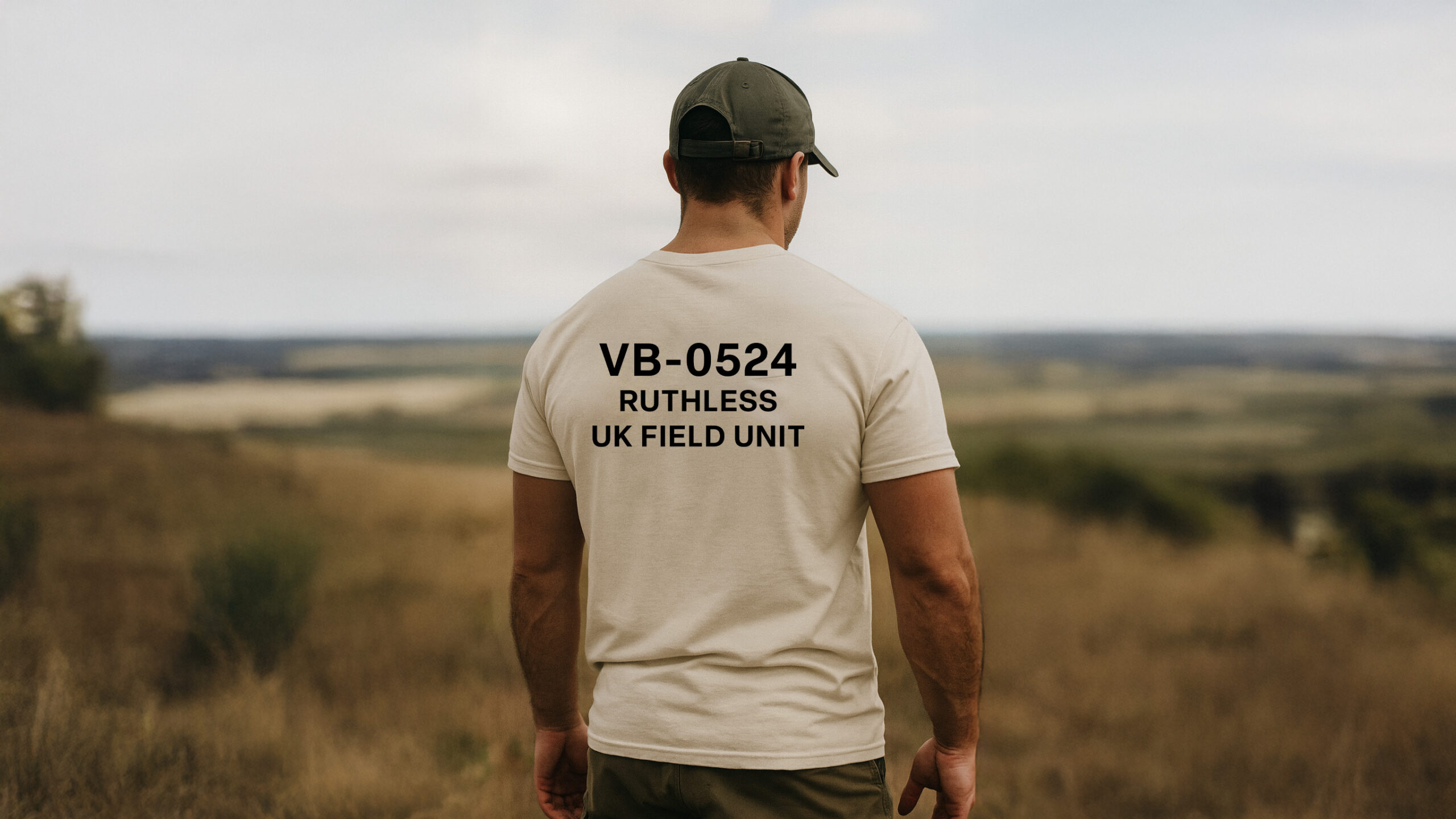 Man standing in open countryside wearing the Varbridge VB-0524 Field Unit natural tee with bold military-style lettering - rugged mission-ready cotton t-shirt built for resilience and intent.