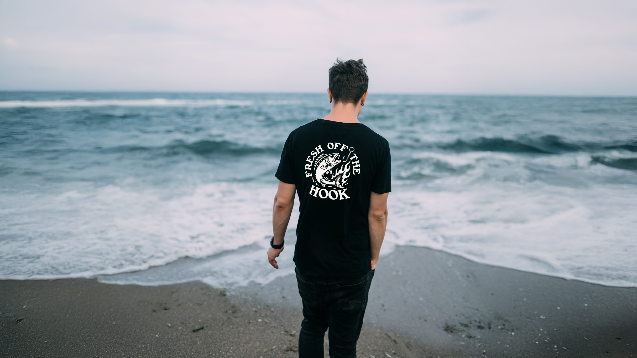 Man standing on a beach facing the ocean wearing the Varbridge “Fresh Off The Hook” black graphic tee with flame-hook fish design — bold fishing-inspired t-shirt for freedom and fearless living.