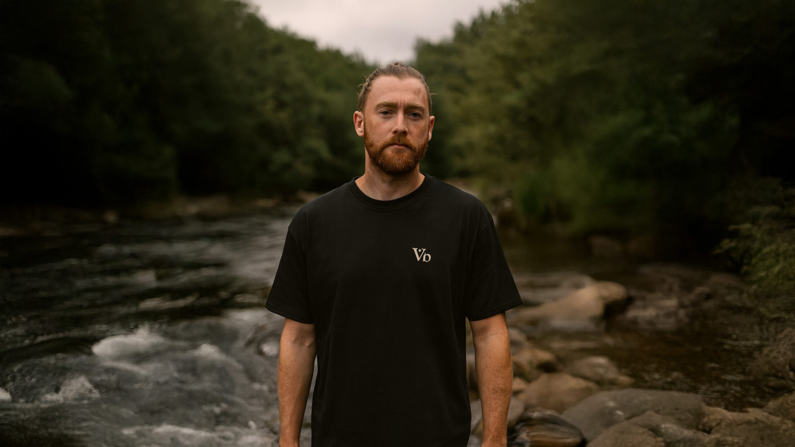 Man wearing the Varbridge “Worn to Work, Built to Last” classic black tee with minimalist Vb chest logo, standing by a rugged riverside in the outdoors.
