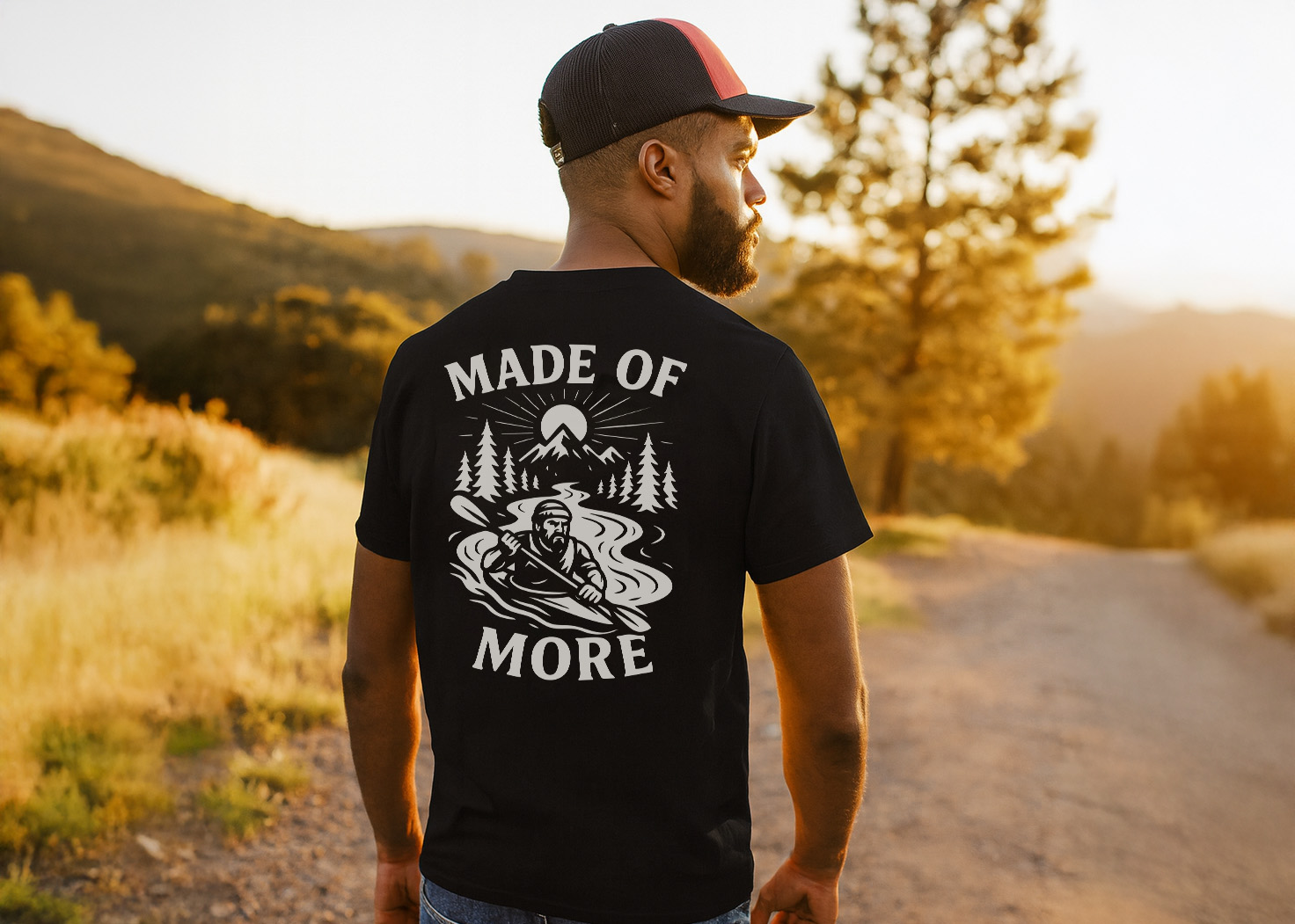 Man wearing a black “Made of More” graphic adventure tee with mountain, river, and kayak design, standing on a sunlit trail in the outdoors.