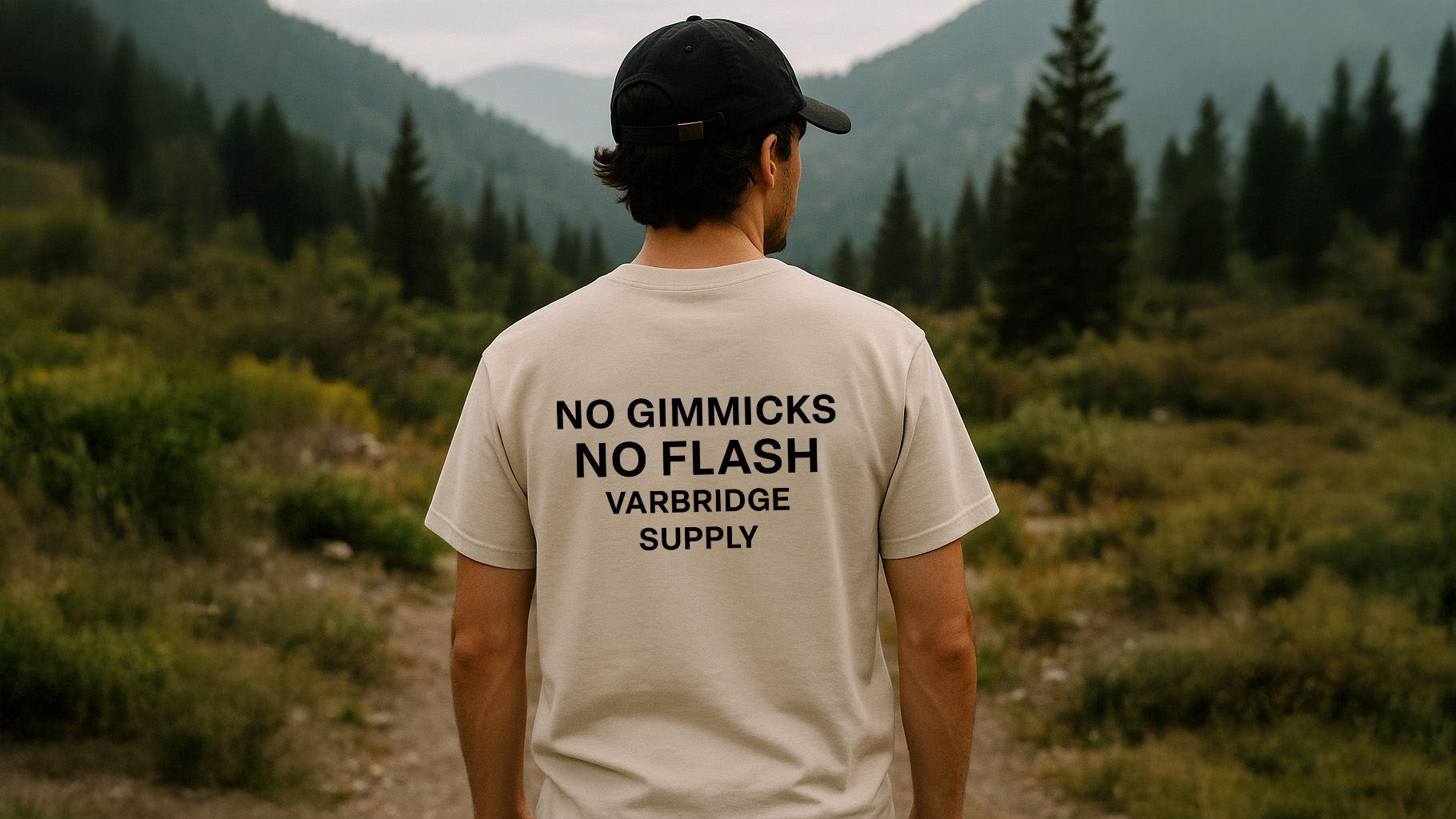 Man standing on a mountain trail wearing the Varbridge “No Gimmicks, No Flash” natural tee with bold black mission-style lettering — rugged minimalist t-shirt built with purpose and intent.