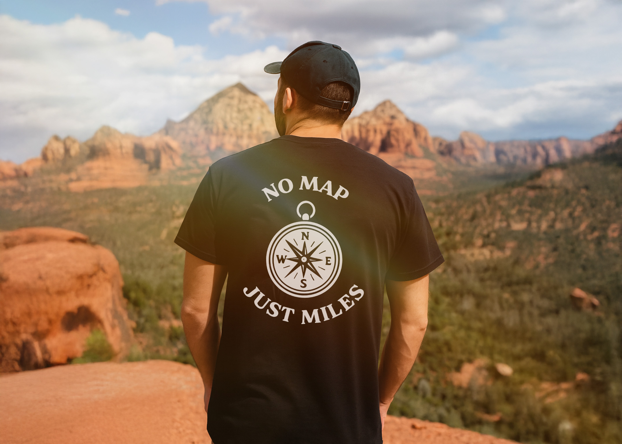 Man wearing a black “No Map, Just Miles” compass graphic tee, standing on a rocky overlook with red mountains and forest in the background.