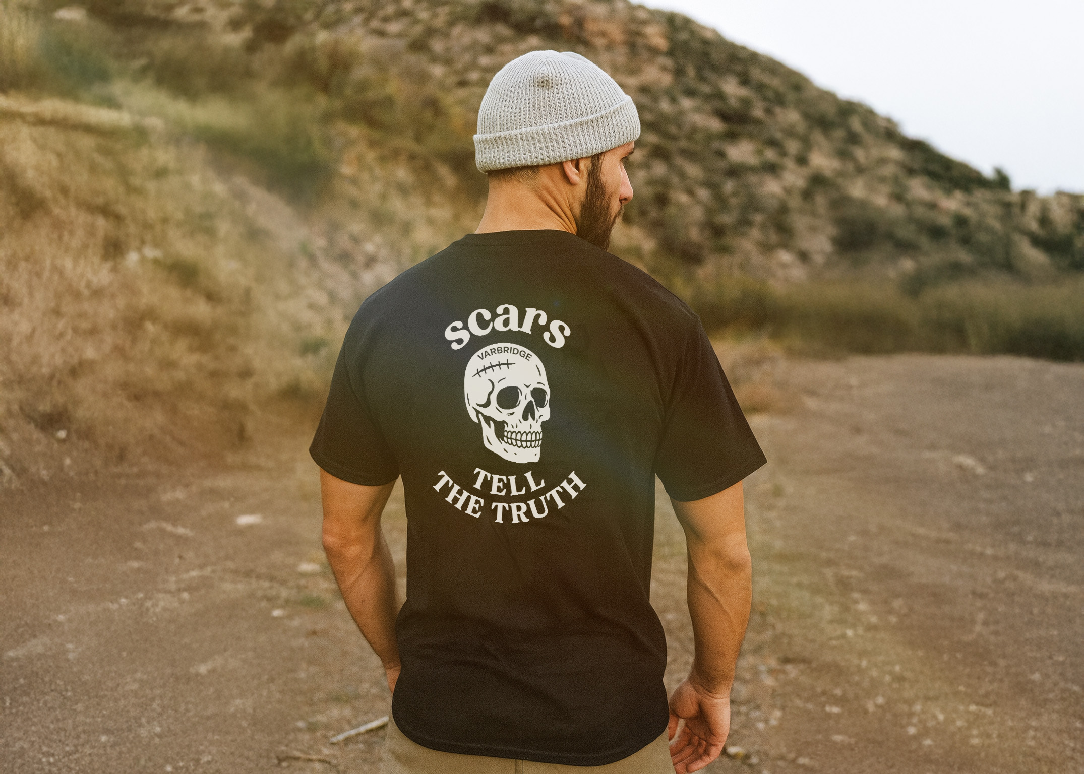 Man wearing a black “Scars Tell the Truth” skull graphic tee and grey beanie, standing on a dirt trail with rocky hills in the background.