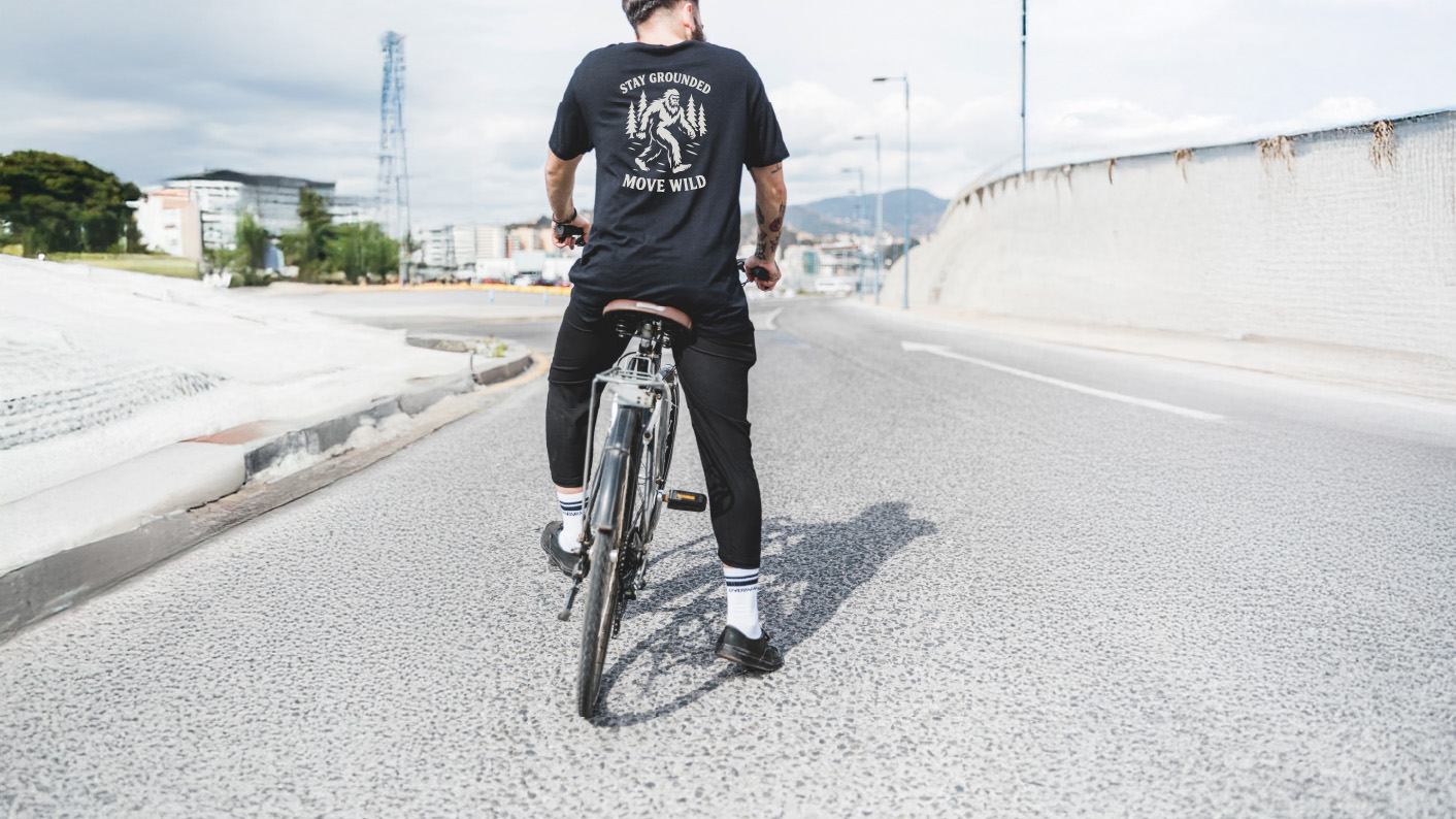 Man cycling through the city wearing the Varbridge “Stay Grounded, Move Wild” black graphic tee with Sasquatch and forest design — adventure-inspired t-shirt blending grit with untamed spirit.