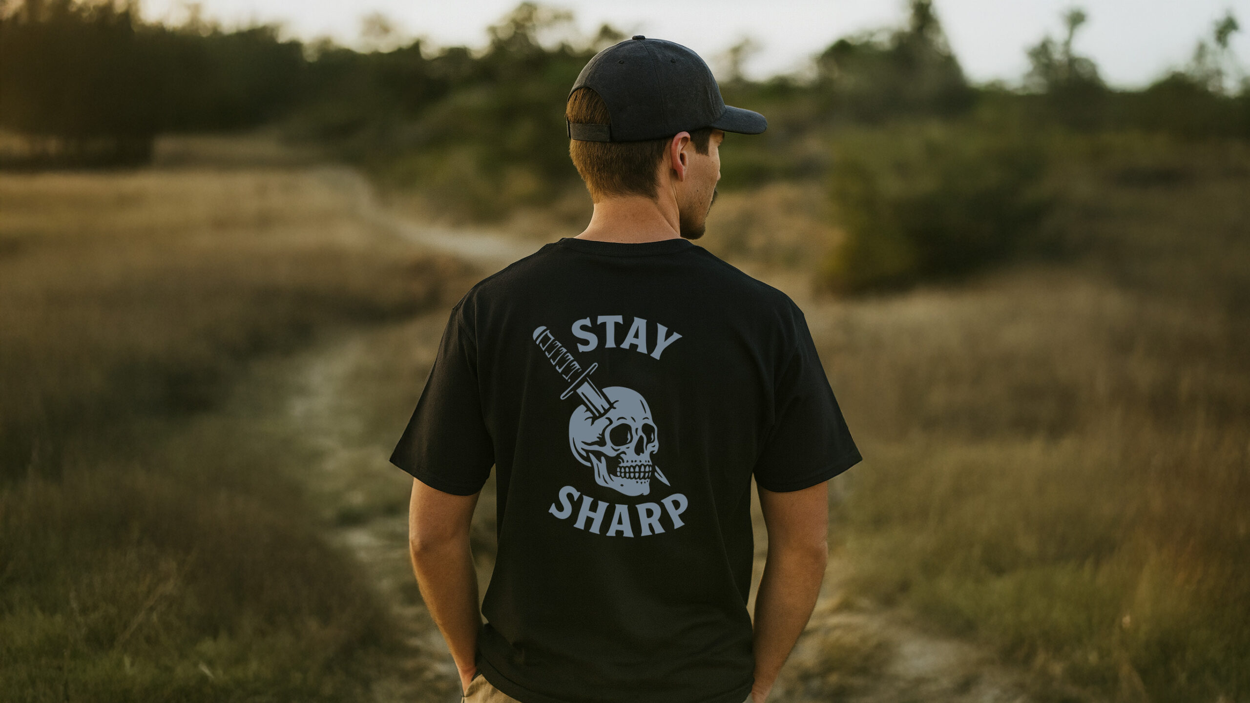 Man walking a dirt trail at sunset wearing the Varbridge “Stay Sharp” black graphic tee with skull and dagger design — tough workwear-inspired t-shirt for focus, grit, and precision.
