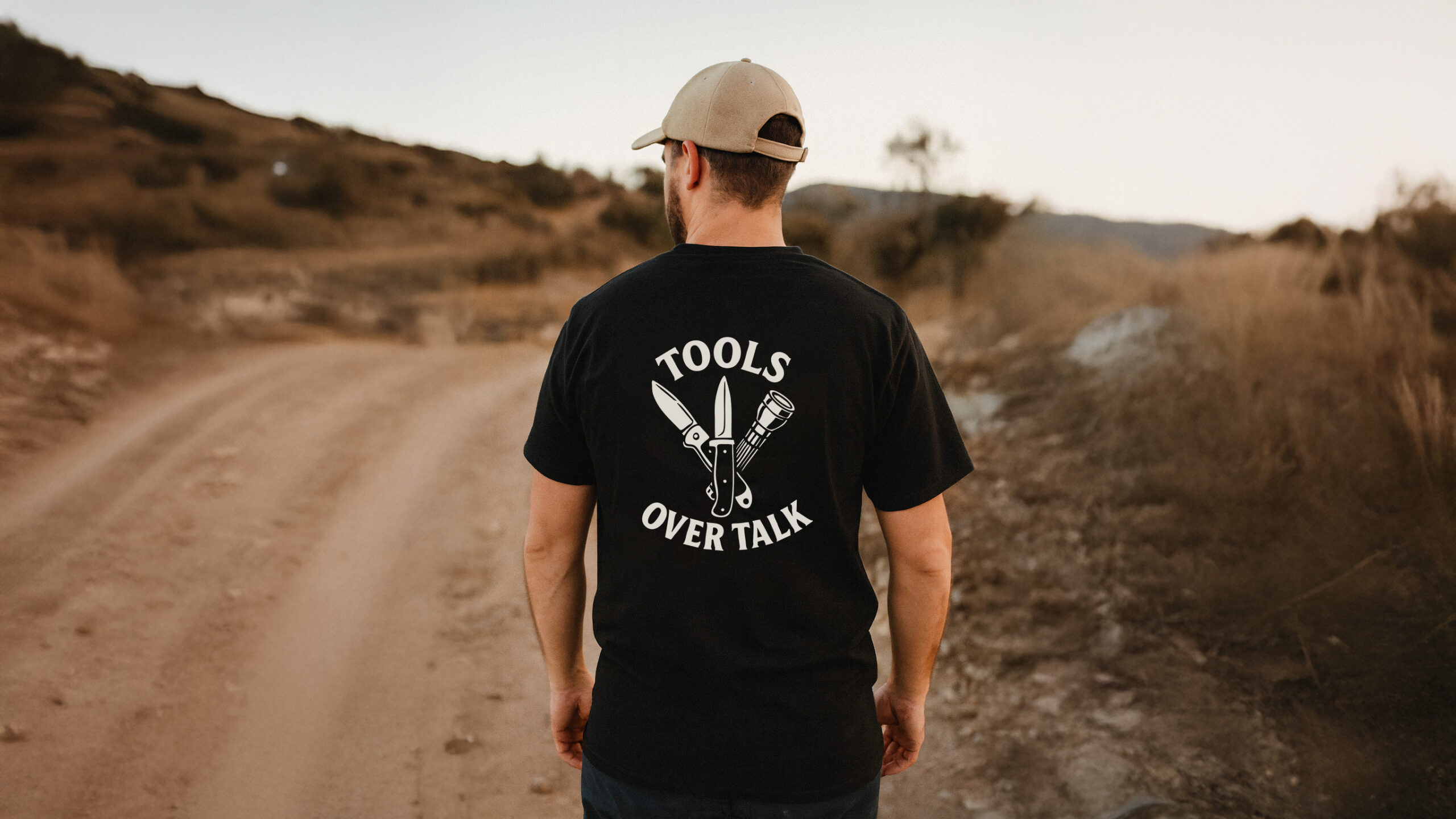 Man walking a rugged dirt road wearing the Varbridge “Tools Over Talk” black graphic tee with crossed knife and torch design - bold workwear-inspired t-shirt built for doers, not talkers.