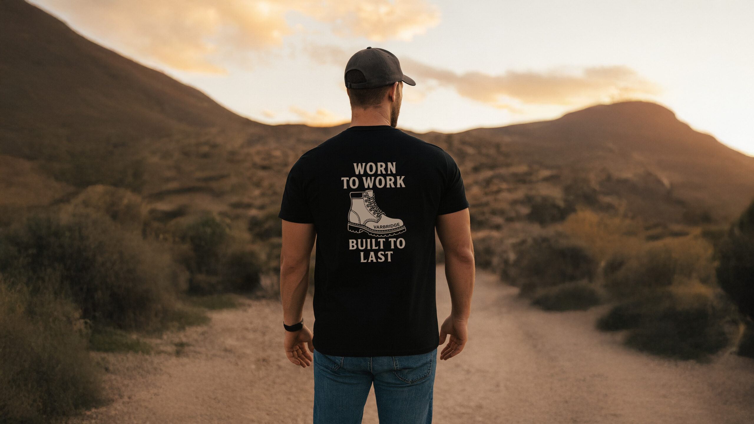 Man wearing the Varbridge “Worn to Work, Built to Last” black graphic tee, standing on a rugged trail at sunset — durable workwear inspired design with bold boot graphic on the back.