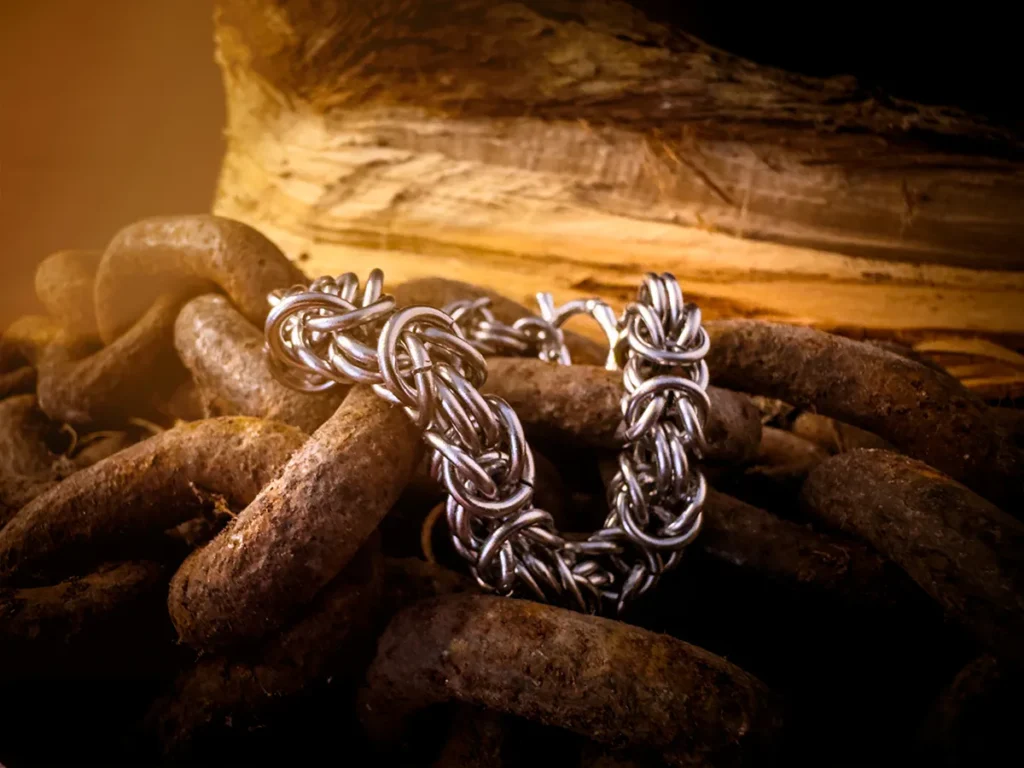 Chainmail-style byzantine men’s stainless steel bracelet looped over heavy rusted chains, with rough-cut wood in the background and warm ambient lighting.