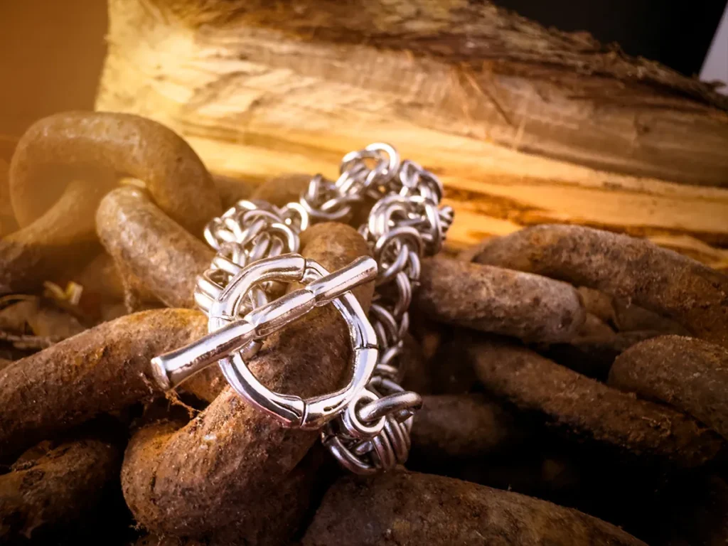 Close-up of a chainmail-style byzantine metal bracelet with a toggle clasp, laid over thick rusted chains with raw wood in the background.
