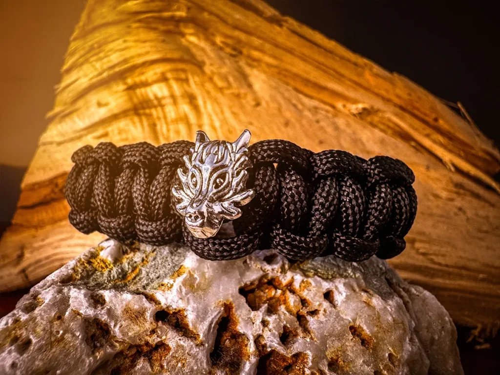 Thick black paracord bracelet with a detailed silver wolf head centerpiece, positioned on rugged quartz with raw wood in the background