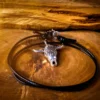Silver longhorn skull pendant on a black adjustable cord, coiled neatly on a polished wood surface with rough-cut timber in the background.