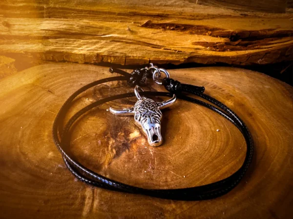 Silver longhorn skull pendant on a black adjustable cord, coiled neatly on a polished wood surface with rough-cut timber in the background.