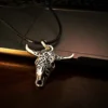 Silver longhorn skull pendant on a black cord, resting on the edge of a dark laptop and wooden surface under low, warm lighting.