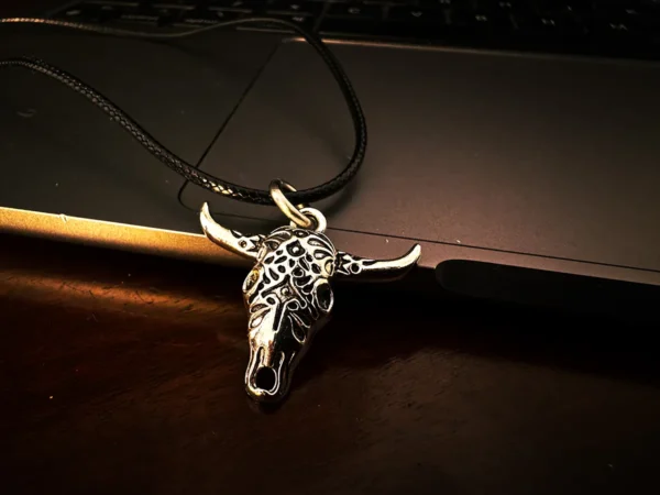 Silver longhorn skull pendant on a black cord, resting on the edge of a dark laptop and wooden surface under low, warm lighting.