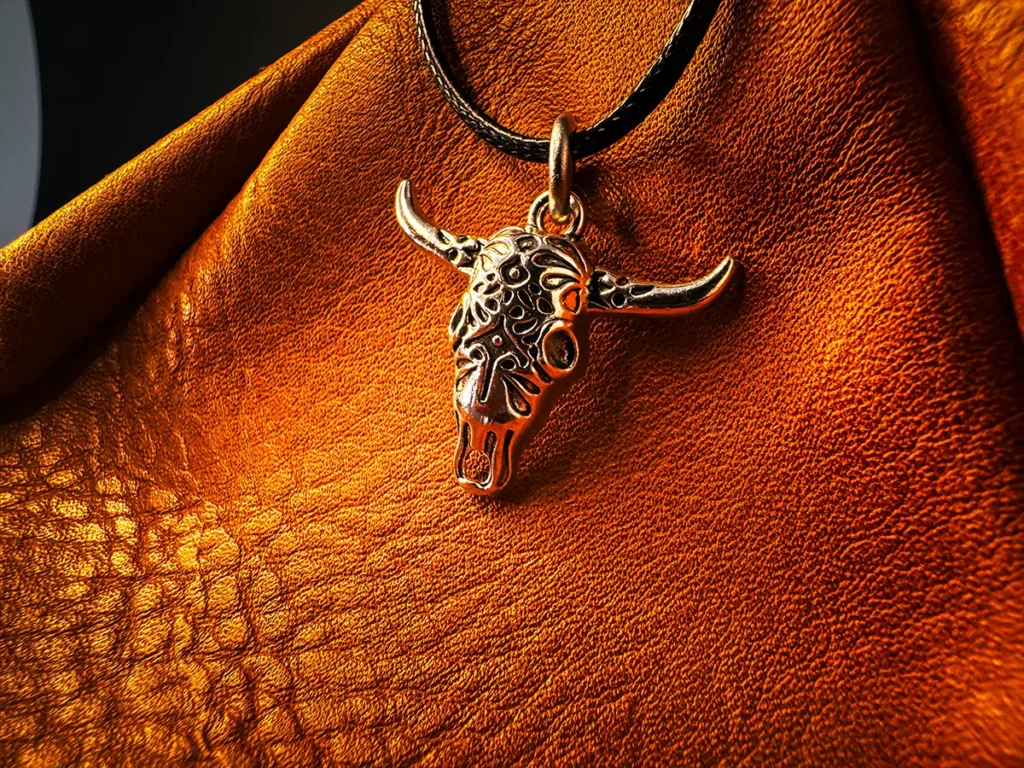 Silver longhorn skull pendant on a black cord, displayed against rich textured brown leather with warm, directional lighting.