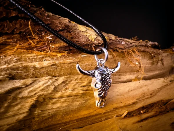 Silver longhorn skull pendant on a black cord, hanging against rough-cut wood with deep grain and dark background contrast.
