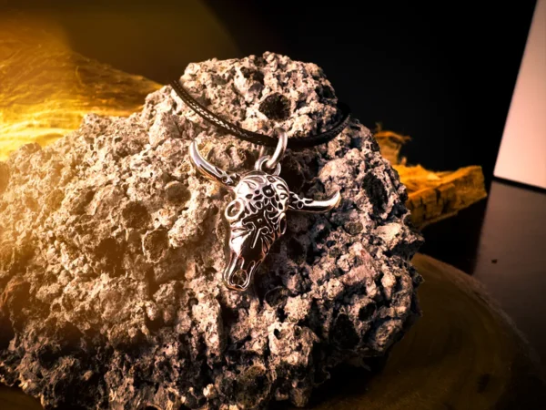 Silver longhorn skull pendant on a black cord, displayed against rough volcanic rock with warm lighting and dark background elements.