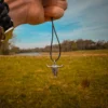 Hand holding a silver bull skull pendant on a black wax cord against a wide-open grassy field, with a river and tree line under a bright, partly cloudy sky- wrist adorned with a rugged black paracord bracelet and metal clasp.