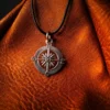 Silver compass pendant on a black cord, displayed against textured brown leather with deep grain and soft, directional lighting.
