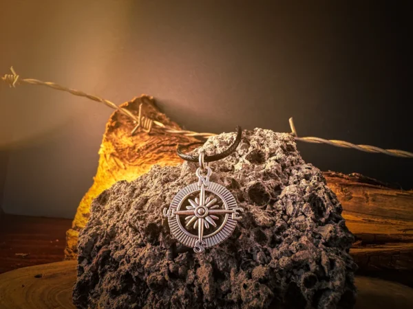 Silver compass pendant on a black cord, displayed on rugged volcanic rock with barbed wire and warm-toned wood in the background.