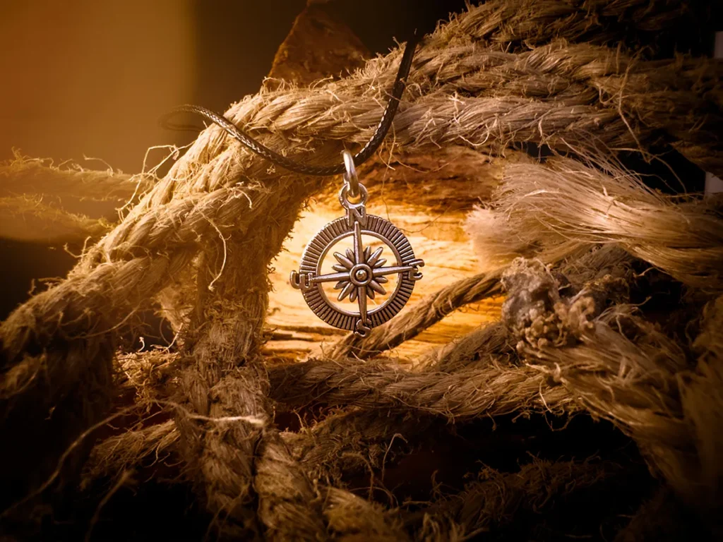 Silver compass pendant on a black cord, hanging among thick, frayed rope and rough-cut wood under warm, moody lighting.
