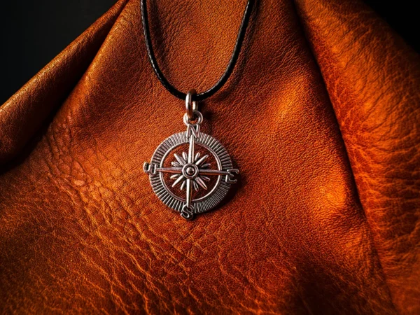 Silver compass pendant on a black cord, displayed against textured brown leather with deep grain and soft, directional lighting.