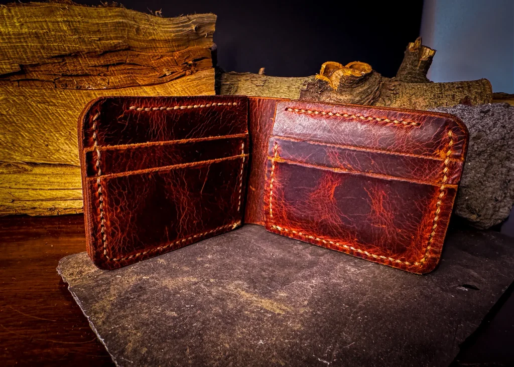 Open handcrafted Full-Grain Leather Wallet showing interior card slots, with rich reddish tones and contrast stitching, placed on a slate surface beside stacked logs and rough stone.