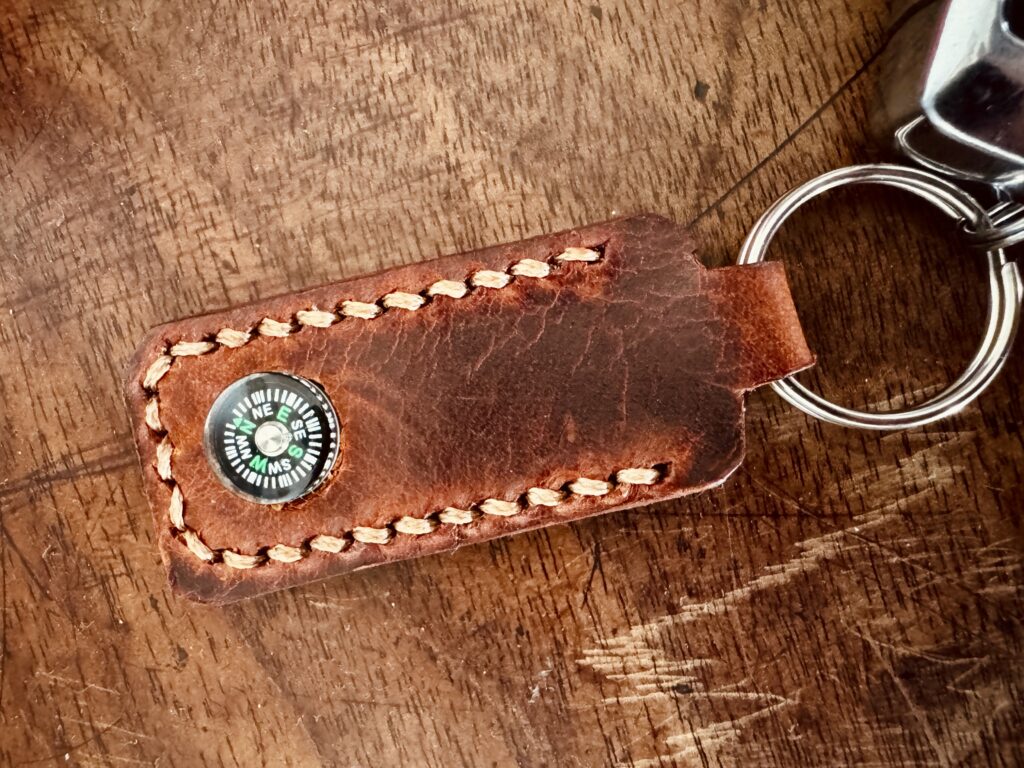 Brown leather keychain with hand-stitched edges and built-in mini compass, attached to a metal keyring, placed on a distressed wooden surface.