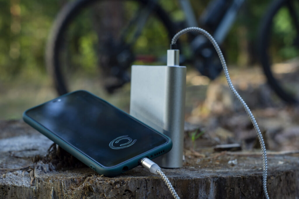 Portable power bank charging a smartphone outdoors with a bicycle in the background – essential tech gift for outdoor enthusiasts and adventurers