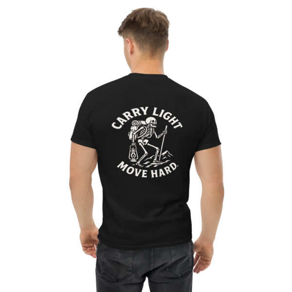 Man wearing a black t-shirt with a white graphic on the back showing a skeleton hiker with a backpack, lantern, and walking stick standing on rocky ground, surrounded by the phrase “CARRY LIGHT MOVE HARD.”