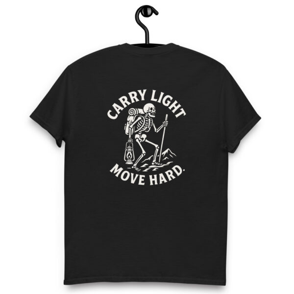 Black t-shirt hanging on a hook with a white graphic on the back showing a skeleton hiker carrying a backpack, lantern, and trekking pole, surrounded by the phrase “CARRY LIGHT MOVE HARD.”