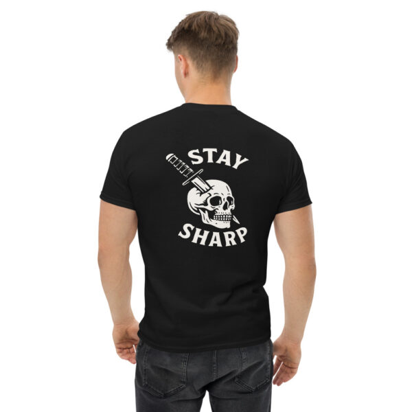 Man wearing a black “Stay Sharp” t-shirt featuring a white graphic of a skull with a tactical knife driven through it, with bold curved text above and below the illustration