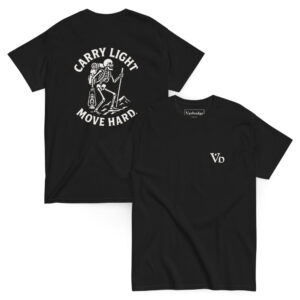 Black t-shirt with a bold white graphic on the back showing a skeleton hiker with a backpack and trekking pole standing on a mountain, accompanied by the phrase “CARRY LIGHT, MOVE HARD.” Front features a minimalist “Vb” logo on the chest.